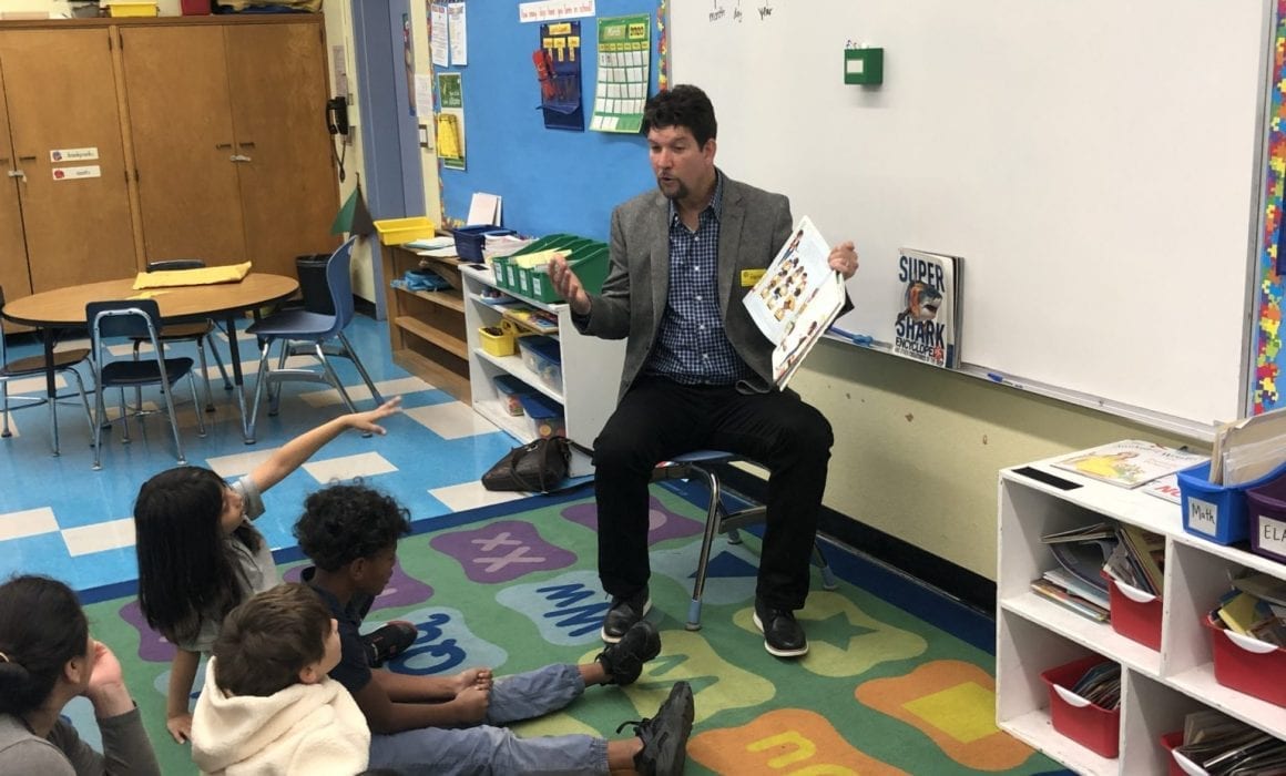 David Goldberg sits in a classroom reading a book to a group of students