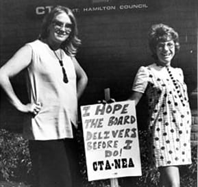 Two pregnant woman stand by a sign that reads I hope the board delivers before I do