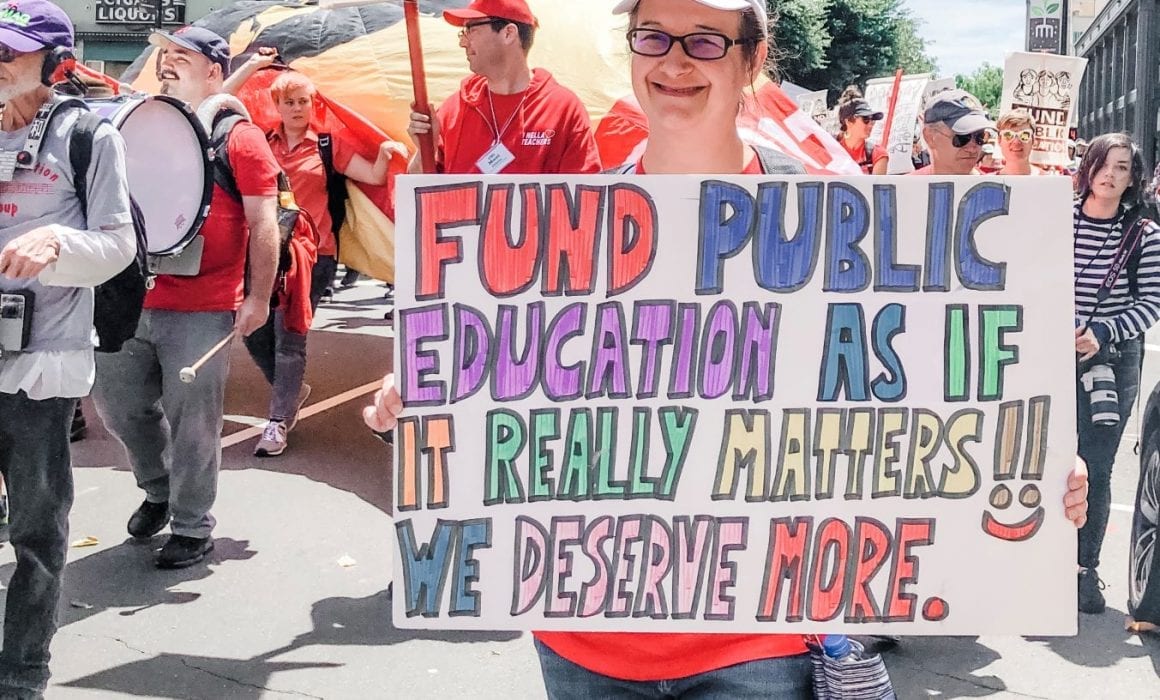 Woman in Red holds sign: Fund Public Education as it if it really matters! We deserve more.