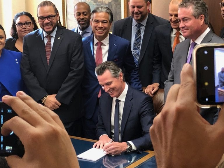 Governor Newsom sits at desk and signs paper as crowd stands around him smiling