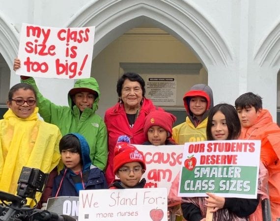 Students stand with Dolores Huerta in raincoats and holding Red For Ed signs
