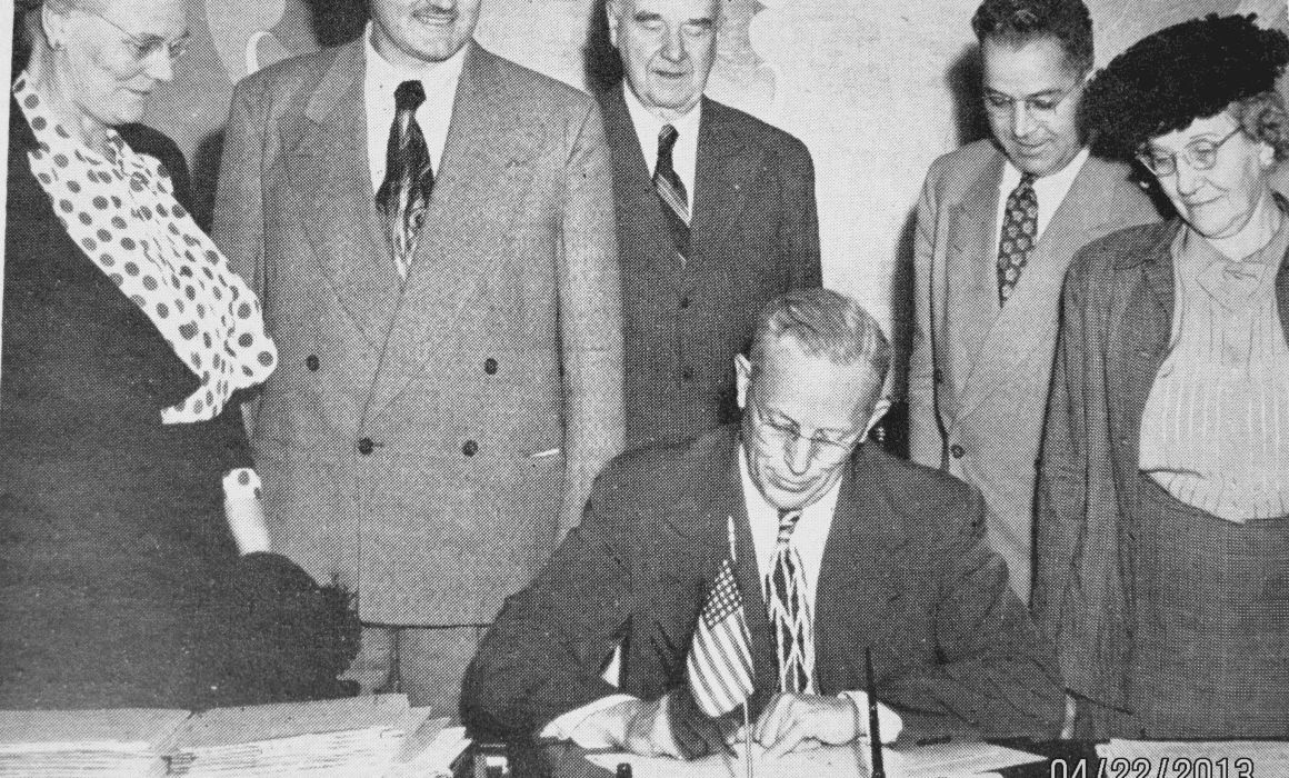 Gov Earl Warren sits at desk signs a piece of legislation with people standing around him