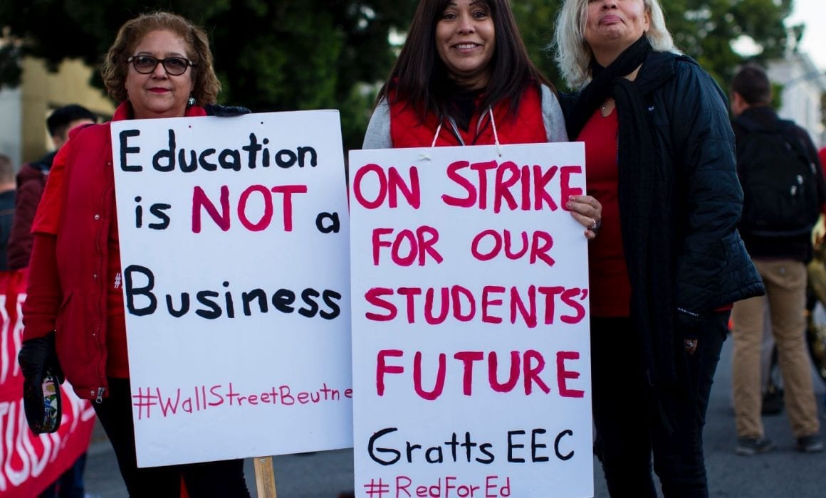 Three women stand together wearing red, two hold signs that read: Education is Not a Business #WallStreetBeutner and On Strike for Our Students Future Gratts EEC #RedForEd