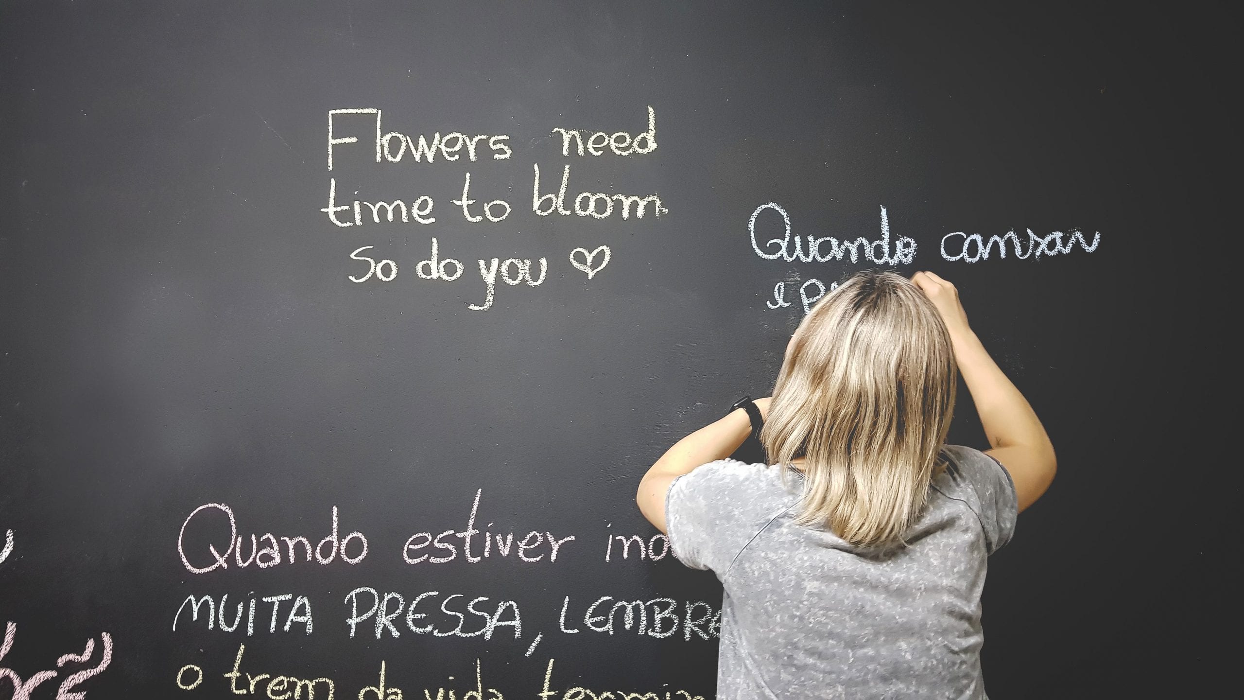 Woman writes on chalkboard: Flowers need time to bloom, so do you.