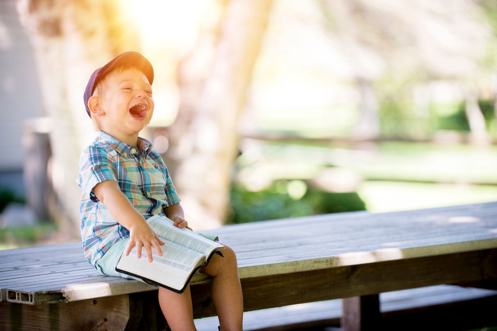 child wearing plaid shirt and hat sitting on bench outside holding books laughs
