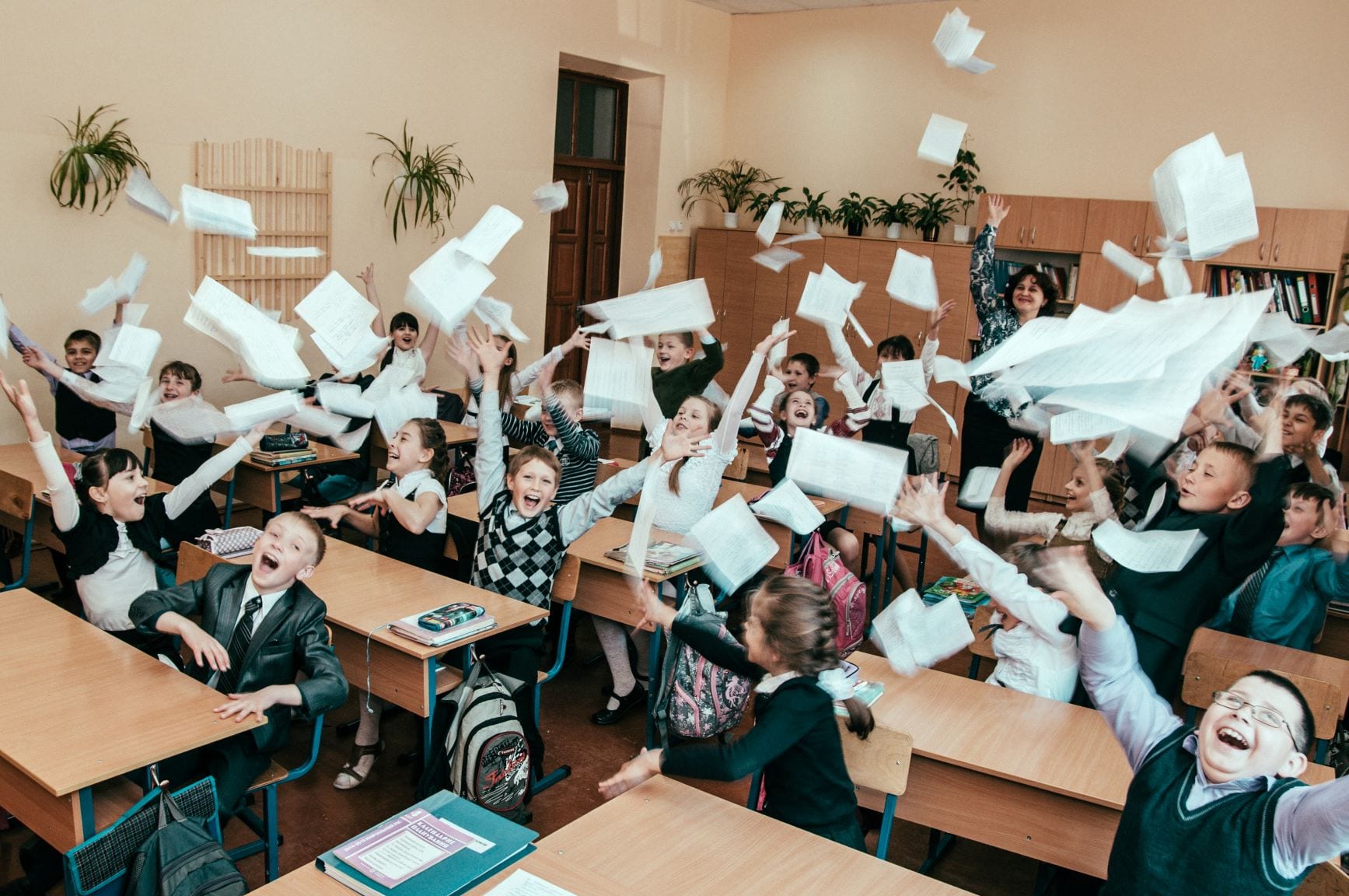 classroom of students laugh and throws papers into air with teacher