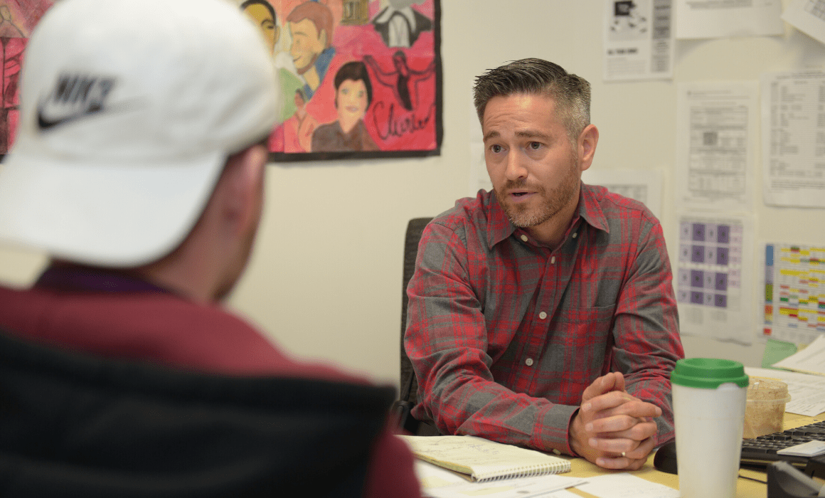 Educator sits at desk and talks to student wearing a baseball hat.