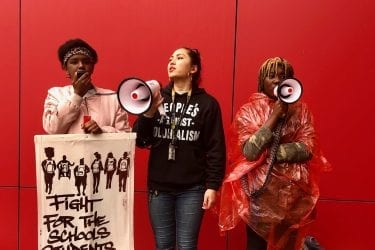 Three women stand in front of red wall holding signs and bullhorns