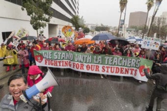 People standing outside in the rain holding a banner that says we stand with LA teachers fighting for our students a person standing in front of them chanting in a bullhorn a