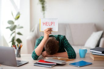 Stock photo of despairing student at desk with laptop holding a sign that says "Help"