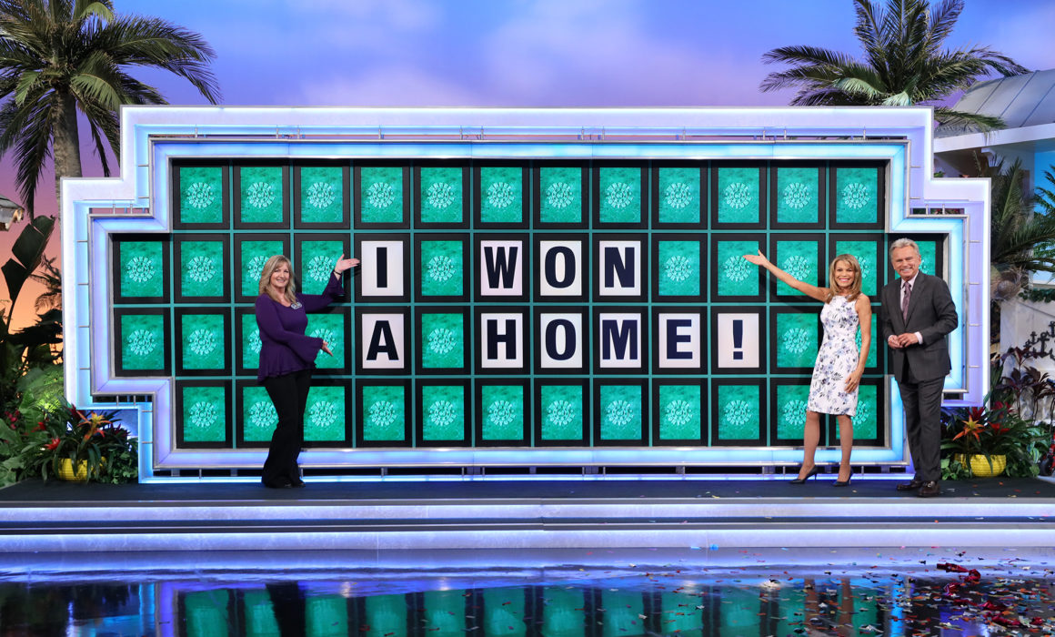 Laura Trammell with "Wheel of Fortune" hosts Pat Sajak and Vanna White in front of big board with words: "I won a home!"