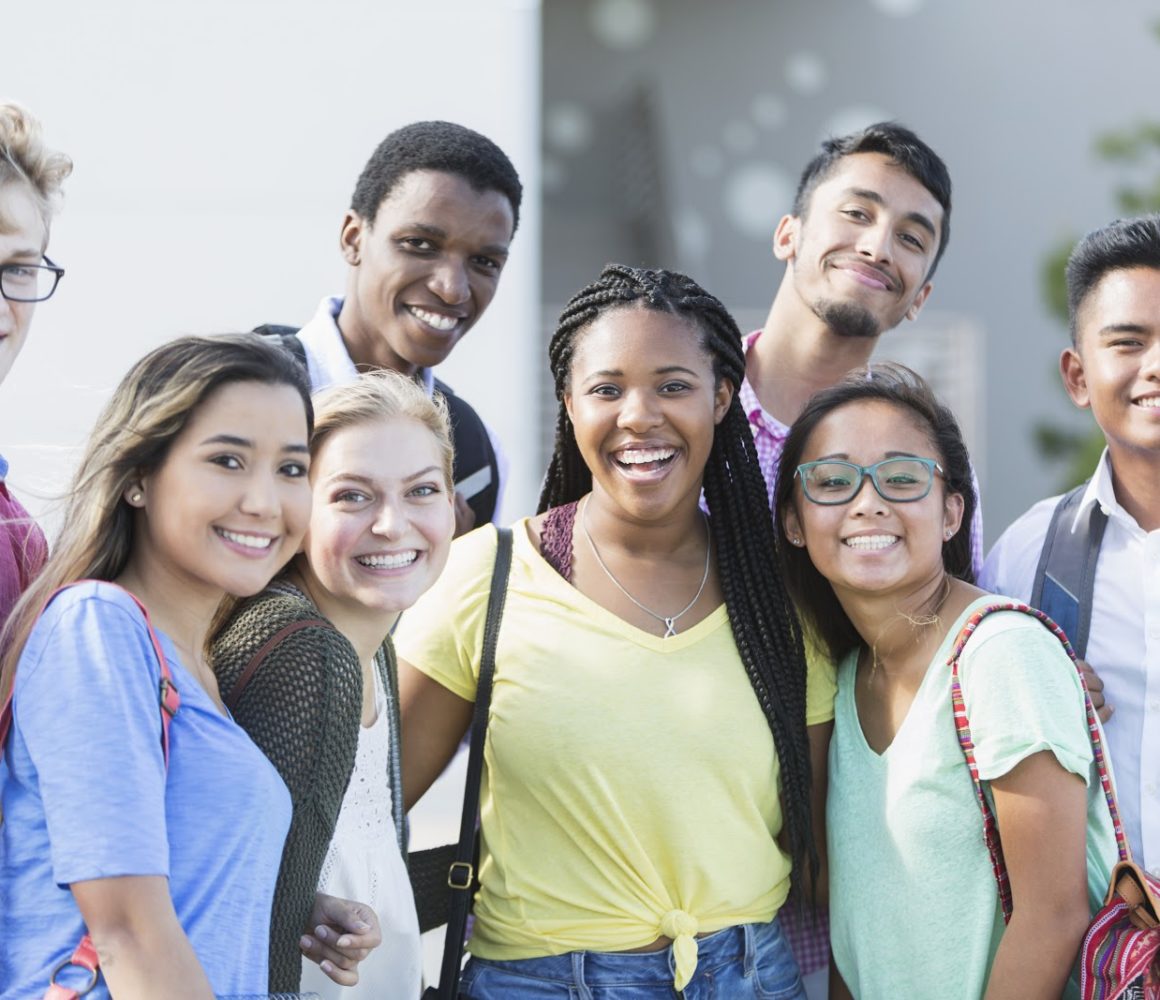 Community Schools - A group of eight multi-ethnic teenagers, 17 and 18 years old, carrying book bags, standing together outside a school building. They are high school seniors