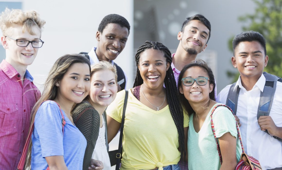 Community Schools - A group of eight multi-ethnic teenagers, 17 and 18 years old, carrying book bags, standing together outside a school building. They are high school seniors