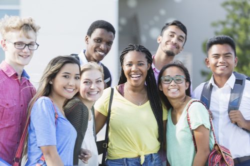 Community Schools - A group of eight multi-ethnic teenagers, 17 and 18 years old, carrying book bags, standing together outside a school building. They are high school seniors