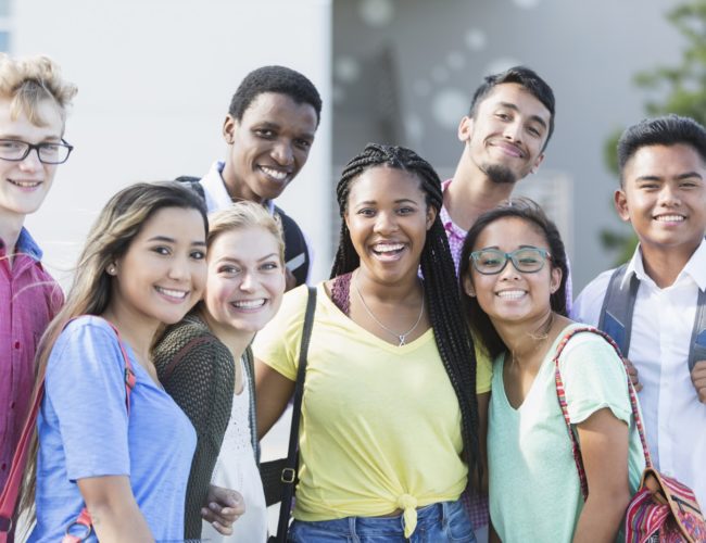 Community Schools - A group of eight multi-ethnic teenagers, 17 and 18 years old, carrying book bags, standing together outside a school building. They are high school seniors