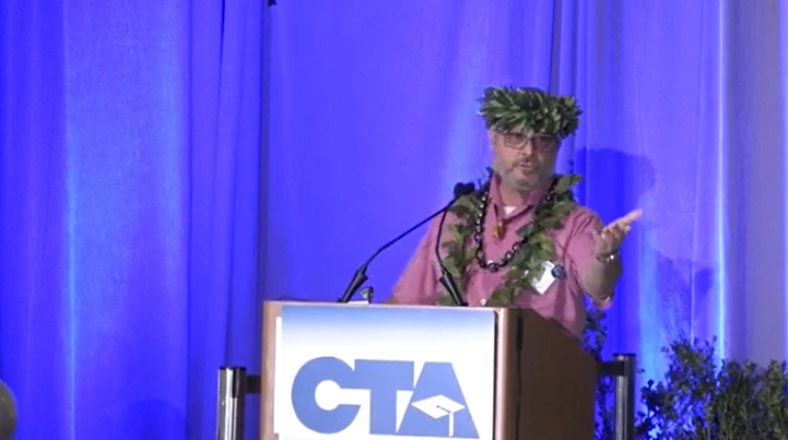 Man with a green garland speaking at a podium against a blue background