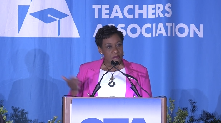 Woman in a pink blazer speaking at a podium against a blue background