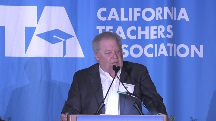 Man in a suit speaking at a podium against a blue background