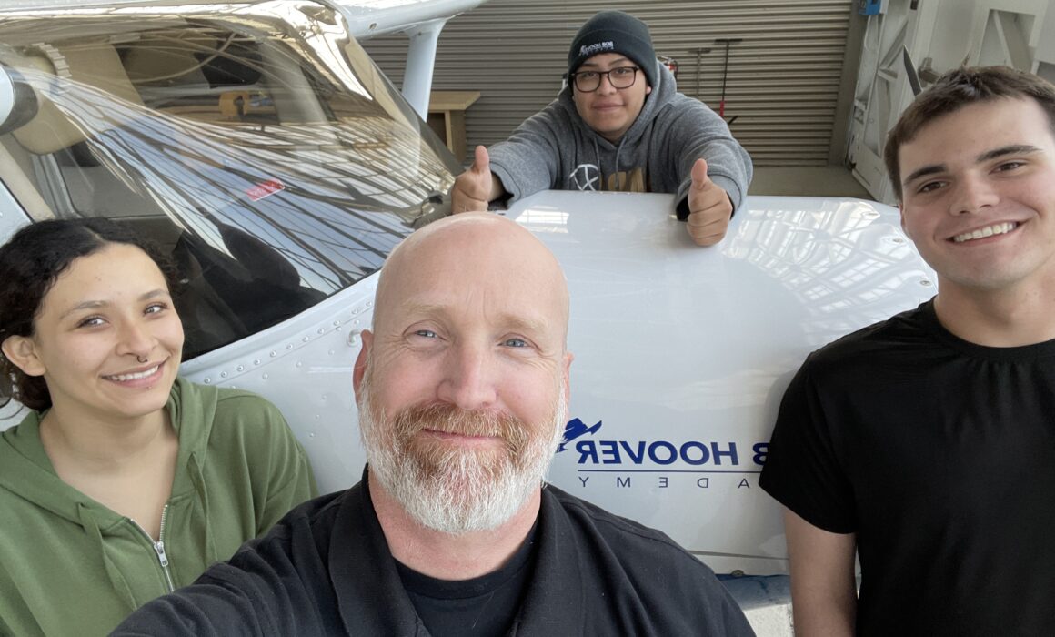 A photo of an educator and three of his students taking a photograph in front of an airplane.