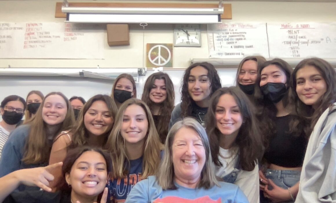 A teacher surrounded by her students pose for a selfie photograph inside a classroom.