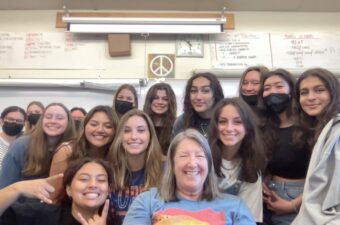 A teacher surrounded by her students pose for a selfie photograph inside a classroom.