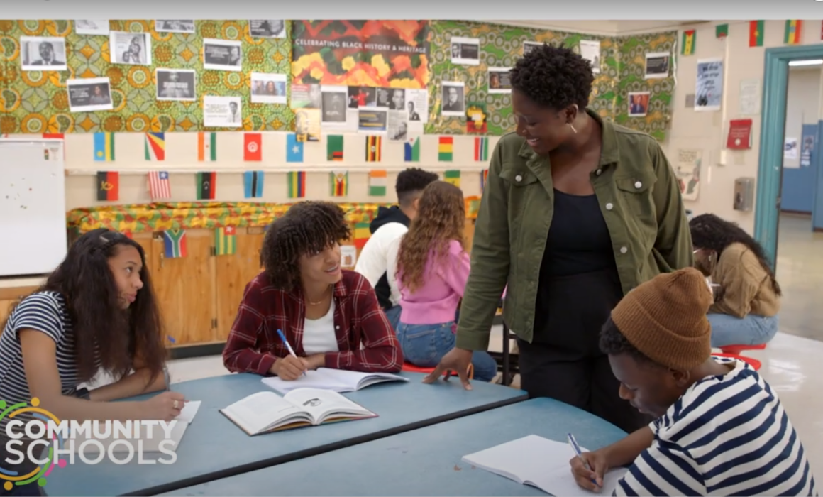 An image of a classroom with students sitting and writing while an adult educator smiles and looks on.