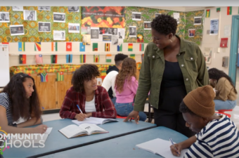 An image of a classroom with students sitting and writing while an adult educator smiles and looks on.