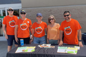 Five teachers posed for a photograph in front of a table. The table cloth displays the following: an orange with the word recall on it and the words: Protect Public Education.