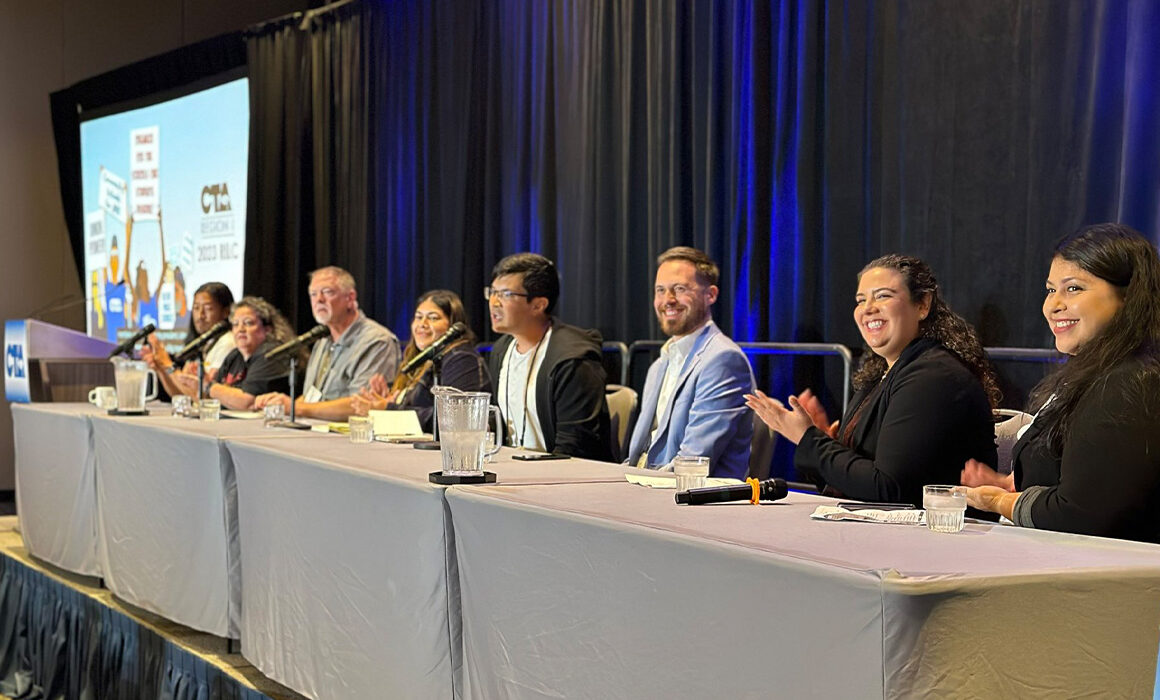 Eight people sit at desks on a stage while smiling and clapping.