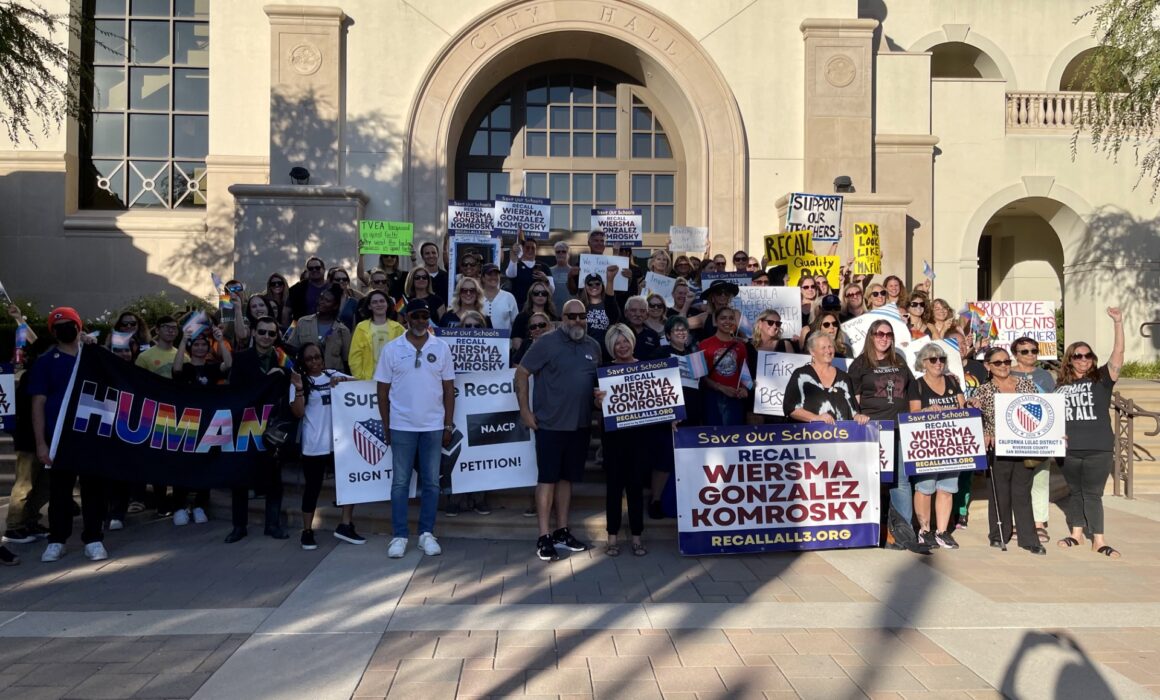 A large group of people pose for a photograph in front of a school holding up protest signs.