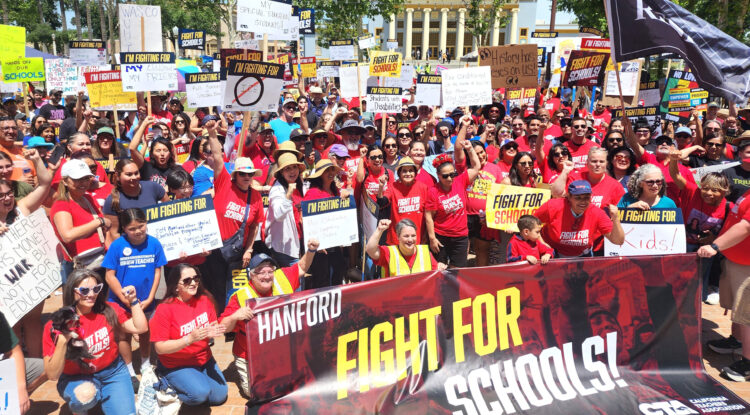 Photo of a large crowd of people holding protest signs in front of a large banner that says "Fight For Schools".